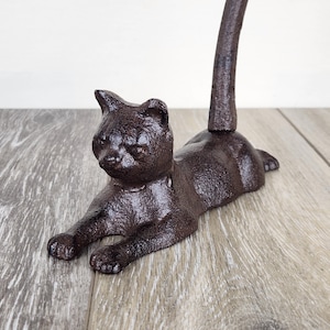 May include: A brown cast iron cat figurine in a relaxed pose. The cat is lying down with its front paws extended and tail curved upwards. The figurine has a textured surface and is likely a decorative item.