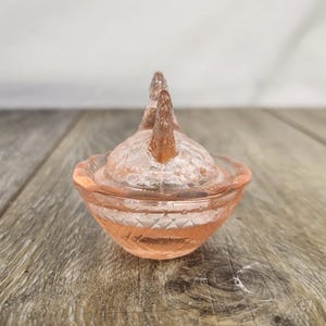 May include: A small, antique, pink glass trinket dish with a lid. The dish has a textured surface and a decorative design on the sides. The lid features two raised, decorative elements. The dish is sitting on a wooden surface.