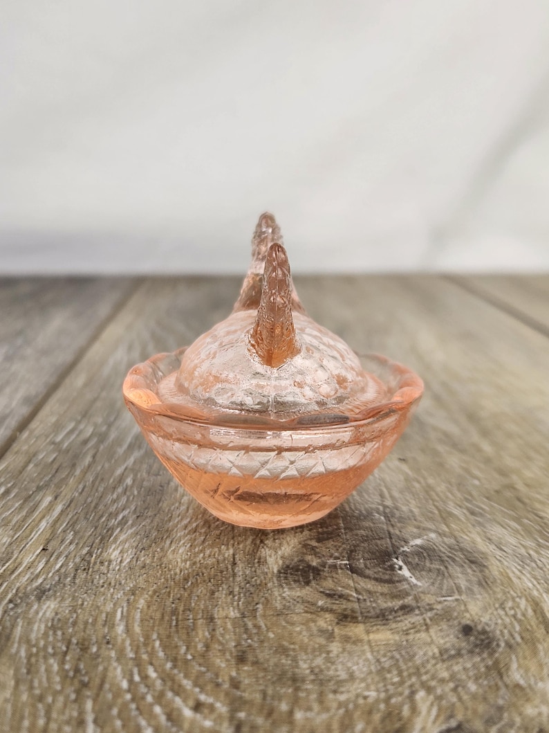 May include: A small, antique, pink glass trinket dish with a lid. The dish has a textured surface and a decorative design on the sides. The lid features two raised, decorative elements. The dish is sitting on a wooden surface.