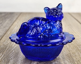 Peut inclure: Une figurine de chat en verre bleu cobalt, posée sur un plat assorti. Le chat est sculpté en position de repos, avec une texture de fourrure détaillée. Le plat a un bord festonné et un design côtelé. Boîte à bijoux décorative.