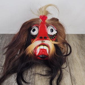 Pu&ograve; includere: Una maschera artigianale con un viso rosso, denti bianchi e lunghi baffi gialli. La maschera ha grandi occhi bianchi ed &egrave; circondata da capelli castani e neri. Un ciuffo di capelli &egrave; sulla parte superiore della maschera.