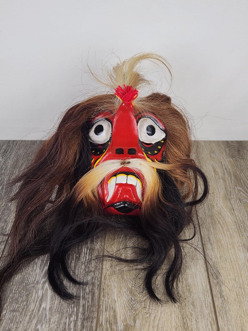Pu&ograve; includere: Una maschera artigianale con un viso rosso, denti bianchi e lunghi baffi gialli. La maschera ha grandi occhi bianchi ed &egrave; circondata da capelli castani e neri. Un ciuffo di capelli &egrave; sulla parte superiore della maschera.