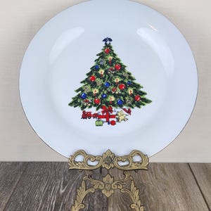 May include: White ceramic plate with a gold rim, featuring a Christmas tree design with red and blue ornaments, and gifts at the base. The plate is displayed on a decorative gold-colored stand, perfect for holiday decor.