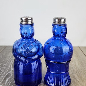 May include: Two cobalt blue glass salt and pepper shakers. The salt shaker is shaped like a woman in a dress with her arms crossed. The pepper shaker is shaped like a man. Both have silver metal lids.