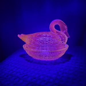 May include: A vintage, pressed glass swan-shaped trinket dish. The dish is a soft, peachy-pink color and features a detailed design on the swan's body and the base. The swan's neck curves gracefully, and the dish sits on a round, decorative base.