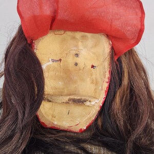 Pu&ograve; includere: Una maschera vintage con un volto bianco sporco e invecchiato, rifiniture rosse e un copricapo in tessuto rosso. Lunghi capelli castano scuro incorniciano il volto. La maschera presenta segni di usura, che suggeriscono et&agrave; e utilizzo.