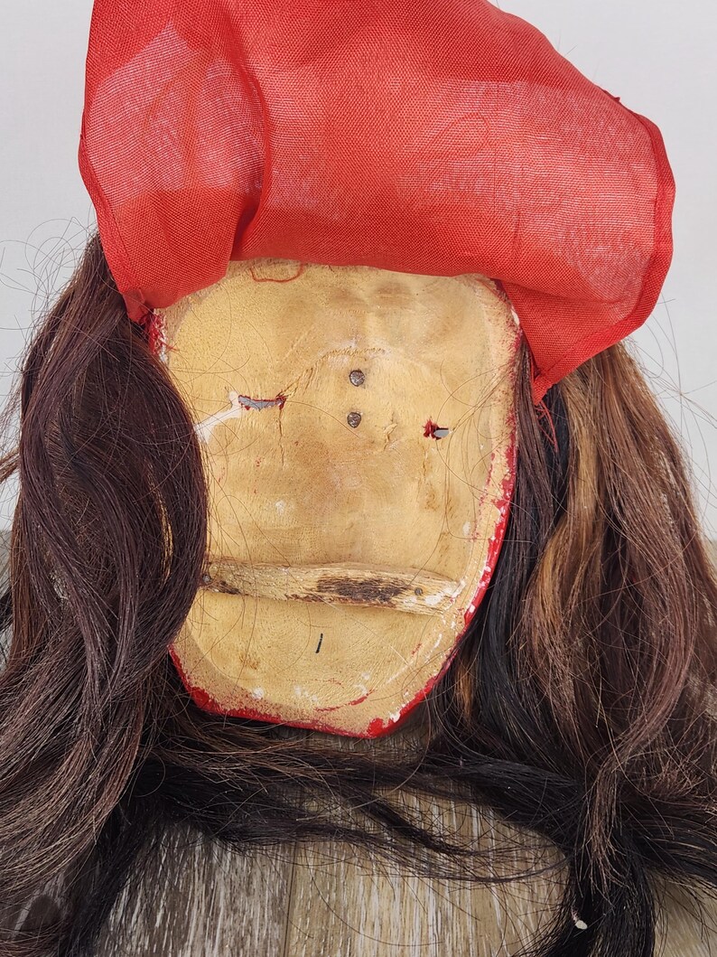 Pu&ograve; includere: Una maschera vintage con un volto bianco sporco e invecchiato, rifiniture rosse e un copricapo in tessuto rosso. Lunghi capelli castano scuro incorniciano il volto. La maschera presenta segni di usura, che suggeriscono et&agrave; e utilizzo.