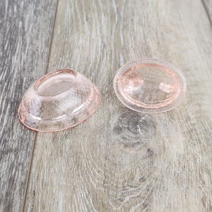 May include: Two pink glass dishes or candle holders on a light wood surface. One is oval with a textured pattern, the other a matching piece with a raised, rounded centre. The items are likely vintage or antique.