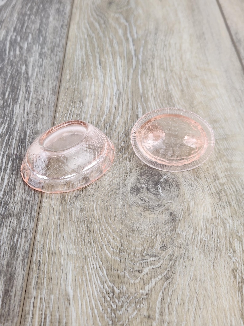 May include: Two pink glass dishes or candle holders on a light wood surface. One is oval with a textured pattern, the other a matching piece with a raised, rounded centre. The items are likely vintage or antique.