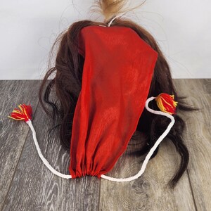 Pu&ograve; includere: Un copricapo in tessuto rosso con fondo arricciato, attaccato a un cordoncino bianco con nappe rosse e gialle. Il copricapo &egrave; circondato da capelli castani e un piccolo ciuffo di capelli biondi.