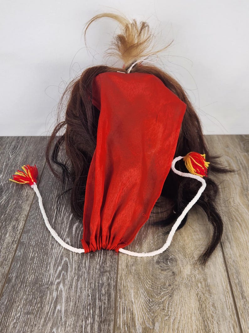 Pu&ograve; includere: Un copricapo in tessuto rosso con fondo arricciato, attaccato a un cordoncino bianco con nappe rosse e gialle. Il copricapo &egrave; circondato da capelli castani e un piccolo ciuffo di capelli biondi.