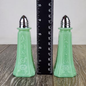 May include: Two vintage jadeite green glass salt and pepper shakers with silver-colored metal tops. The shakers have a floral embossed design and are approximately 5 inches tall.