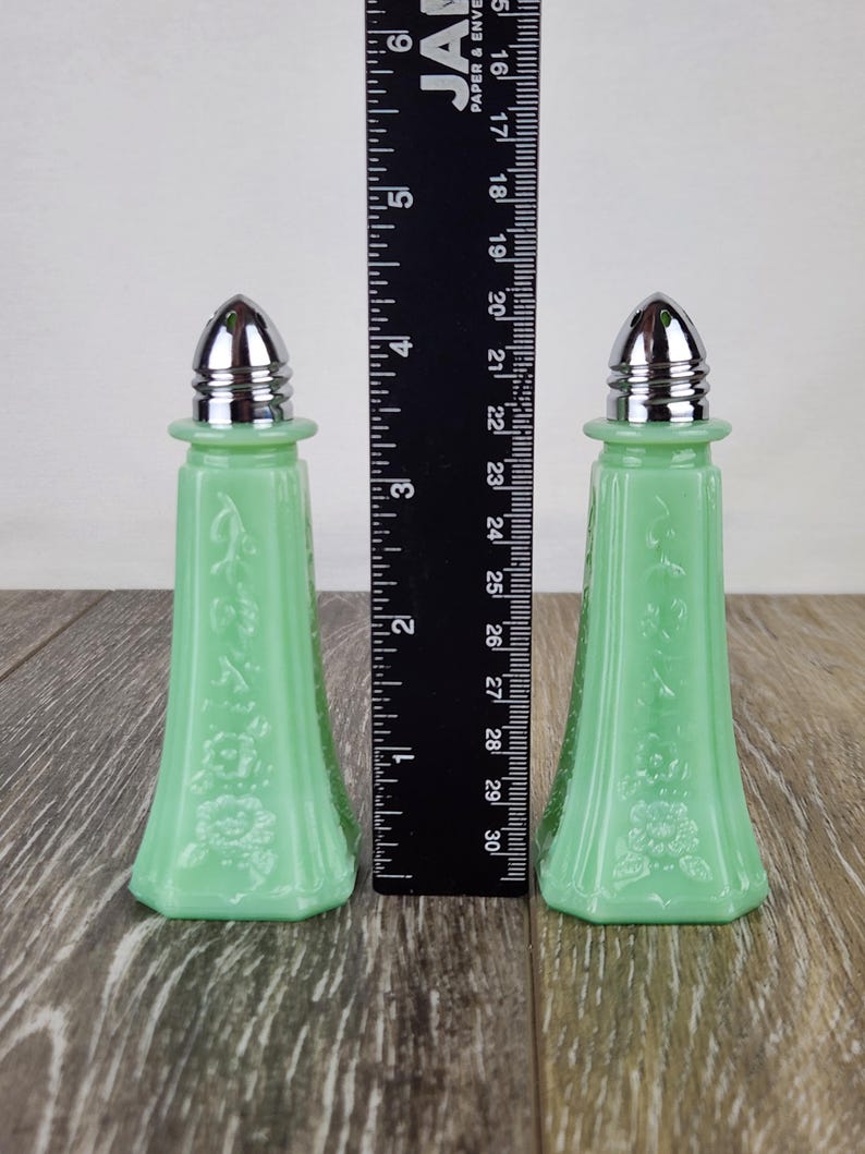 May include: Two vintage jadeite green glass salt and pepper shakers with silver-colored metal tops. The shakers have a floral embossed design and are approximately 5 inches tall.