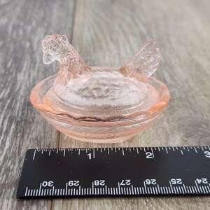 May include: A pink glass hen-shaped dish with a textured surface. The hen has a detailed head and wings, sitting atop a round, textured bowl. The dish is likely used for decorative purposes or as a small trinket holder.