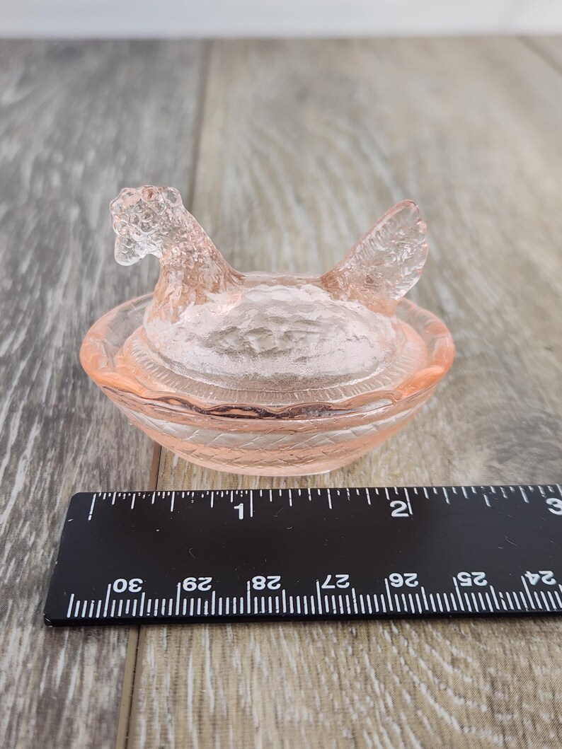 May include: A pink glass hen-shaped dish with a textured surface. The hen has a detailed head and wings, sitting atop a round, textured bowl. The dish is likely used for decorative purposes or as a small trinket holder.