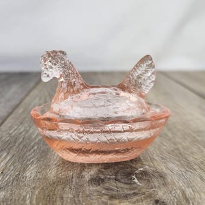 May include: A vintage pink glass hen-on-nest dish. The hen-shaped lid sits atop a round, textured bowl. The glass has a translucent quality, and the dish is displayed on a wooden surface. A decorative piece for the kitchen or dining room.
