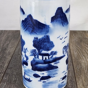 May include: A cylindrical ceramic vase featuring a white base with blue hand-painted landscape illustrations. The design depicts mountains, trees, a small structure, and a boat on water. The vase has a smooth, glossy finish and an open top.