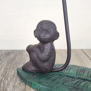May include: A dark brown monkey figurine sitting on a green leaf-shaped base. The monkey is in a seated position with its arms crossed. A black metal rod extends upward from the base.