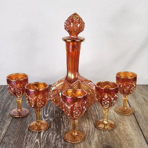 May include: A set of orange iridescent carnival glass consisting of one decanter with a matching stopper and four stemmed glasses. Each piece features a pressed geometric diamond and starburst pattern with a reflective, metallic finish.