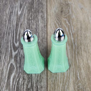 May include: Two vintage, light green salt and pepper shakers with silver-colored metal tops. The glass shakers have a floral design and are displayed on a wooden surface. A classic kitchenware set.