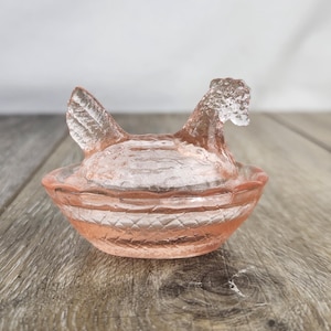 May include: A pink glass hen-shaped dish. The hen has a detailed comb and wattle, and the dish has a ridged design. This decorative piece is likely used for serving or display. The dish is made of glass.