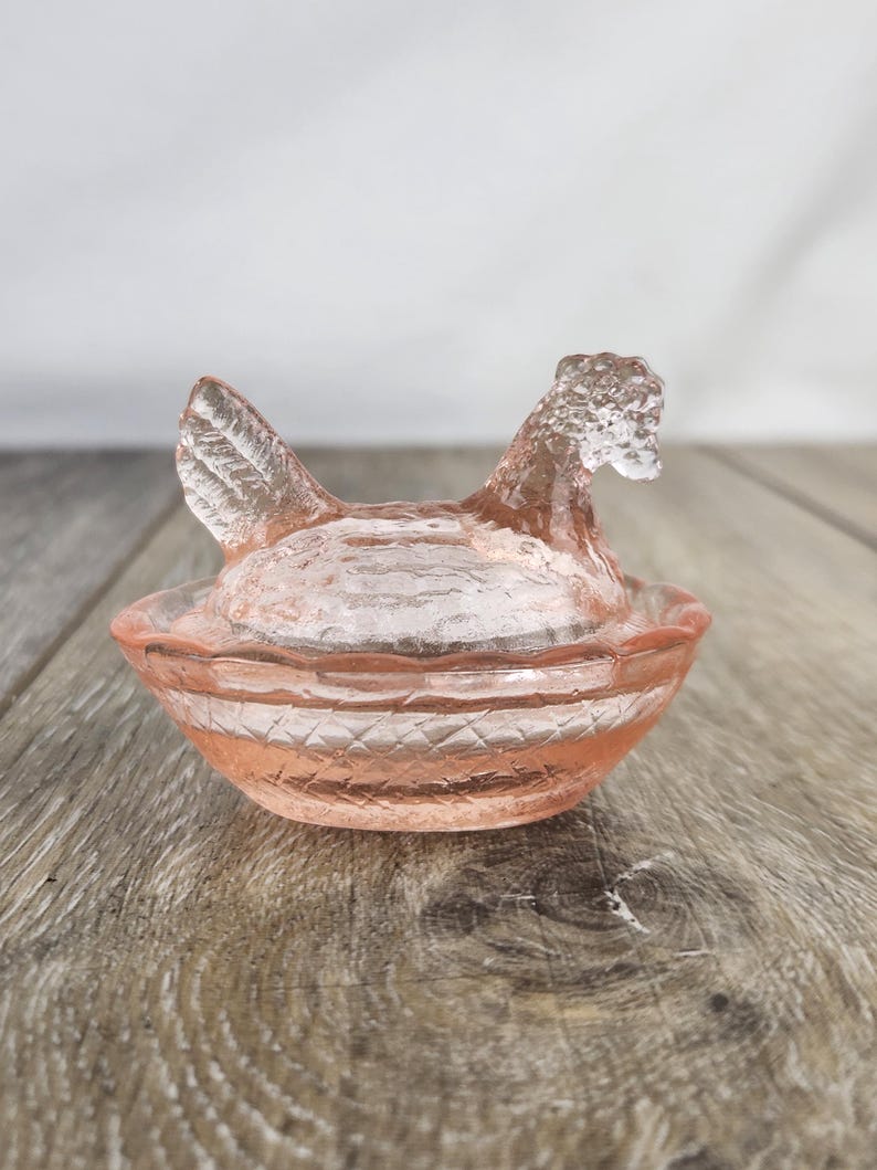 May include: A pink glass hen-shaped dish. The hen has a detailed comb and wattle, and the dish has a ridged design. This decorative piece is likely used for serving or display. The dish is made of glass.