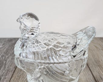 May include: A clear glass hen-shaped trinket box. The lid forms the hen's body and wings, while the base is the lower portion. The glass has a textured, detailed design. The hen is sitting on a wooden surface.