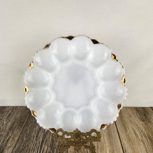 May include: A white, scalloped egg serving plate with gold trim, displayed on a decorative brass stand. The plate has 12 individual egg compartments. The plate is on a wooden surface.
