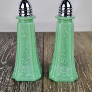 May include: Two matching jadeite green salt and pepper shakers. Each shaker has a flared, octagonal base and a floral design. The tops are silver-colored metal with a ribbed design.