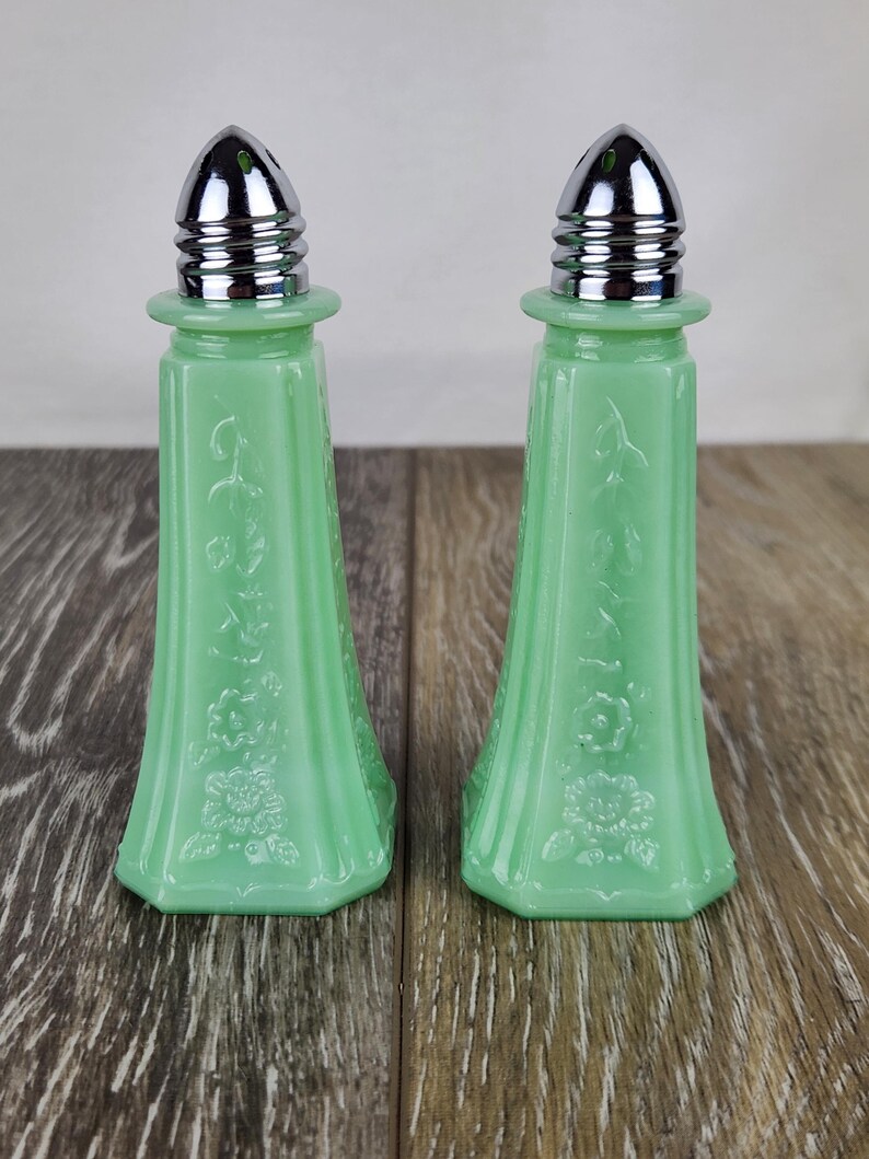 May include: Two matching jadeite green salt and pepper shakers. Each shaker has a flared, octagonal base and a floral design. The tops are silver-colored metal with a ribbed design.
