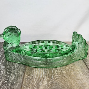 May include: A green glass hen-shaped dish with a detailed design. The dish features a head, body, and tail, with a textured surface. The body has several circular indentations. The dish is made of transparent green glass and sits on a wooden surface.