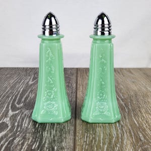 May include: A pair of light green glass salt and pepper shakers with silver-colored metal tops. The shakers have a flared base and are decorated with floral patterns. The shakers are displayed on a wooden surface.