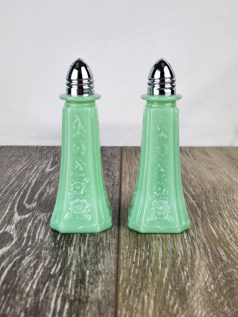 May include: A pair of light green glass salt and pepper shakers with silver-colored metal tops. The shakers have a flared base and are decorated with floral patterns. The shakers are displayed on a wooden surface.