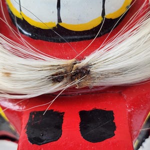 Pu&ograve; includere: Primo piano di una maschera rossa dipinta a mano con denti bianchi, dettagli gialli e dettagli neri. La maschera presenta capelli bianchi e due forme rettangolari nere. La maschera &egrave; probabilmente utilizzata per scopi culturali o teatrali.