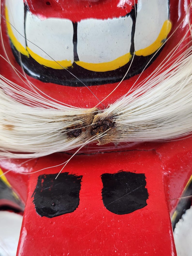 Pu&ograve; includere: Primo piano di una maschera rossa dipinta a mano con denti bianchi, dettagli gialli e dettagli neri. La maschera presenta capelli bianchi e due forme rettangolari nere. La maschera &egrave; probabilmente utilizzata per scopi culturali o teatrali.