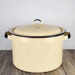 May include: Cream-colored enamel pot with a lid and black trim. The pot has two side handles and a loop handle on the lid. The pot is sitting on a wooden surface.