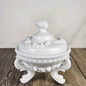 May include: A white, lidded serving dish with a corn-shaped handle. The dish has a detailed, embossed design with floral and shell motifs, and rests on three ornate, curved feet. The overall design is vintage and decorative.