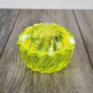 May include: A vibrant, neon-yellow glass trinket box with a lid. The box has a ribbed, spiral design and a starburst pattern on the lid. The box is sitting on a wooden surface.