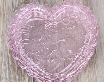 May include: A heart-shaped, pink glass dish with a scalloped edge. The dish features an embossed design of two birds and floral elements. The transparent glass allows light to pass through, highlighting the intricate details of the design.