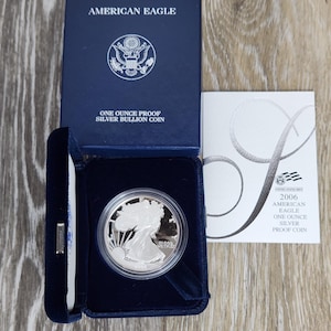 May include: A silver American Eagle one-ounce proof coin in a blue box. The box is open, revealing the coin in a velvet-lined compartment. The box reads "AMERICAN EAGLE" and "ONE OUNCE PROOF SILVER BULLION COIN". A card with the text "2006 AMERICAN EAGLE ONE OUNCE SILVER PROOF COIN" is also visible.