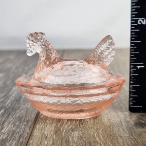 May include: A pink glass hen-shaped dish with a lid. The hen has a textured surface and is sitting in a basket-like bowl. The dish is approximately 10 cm long.