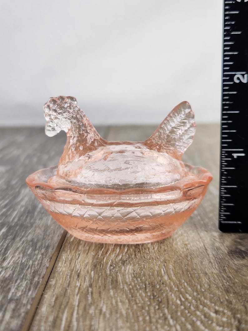 May include: A pink glass hen-shaped dish with a lid. The hen has a textured surface and is sitting in a basket-like bowl. The dish is approximately 10 cm long.