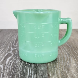 May include: A vintage jadeite green measuring cup with a handle and a pouring spout. The cup has embossed measurement markings for cups and fluid ounces. The cup is made of glass and sits on a wooden surface.