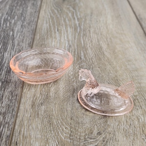 May include: A pink glass trinket dish and a duck-shaped lid. The dish is oval with a textured rim. The lid is a duck with a detailed head and wings. The items are made of translucent pink glass and sit on a wooden surface.
