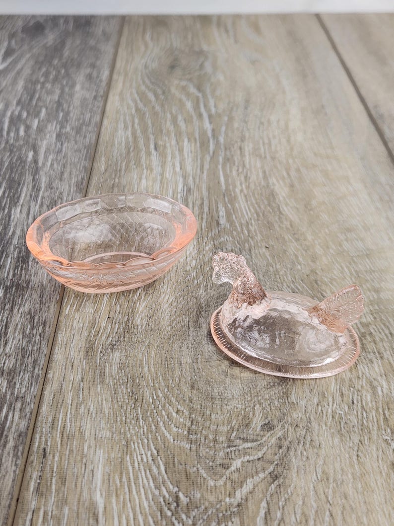 May include: A pink glass trinket dish and a duck-shaped lid. The dish is oval with a textured rim. The lid is a duck with a detailed head and wings. The items are made of translucent pink glass and sit on a wooden surface.