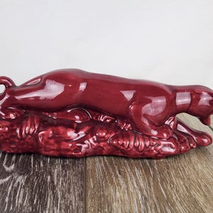 May include: A maroon ceramic panther figurine, lying down on a textured base. The glossy sculpture has a curled tail and an open mouth, showcasing a detailed design. This decorative piece is suitable for home decor.