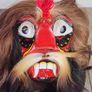 Pu&ograve; includere: Una maschera vibrante e artigianale con un'espressione feroce. La maschera presenta un volto rosso con occhi bianchi, lunghi baffi bianchi e marroni e una bocca rossa con denti bianchi. Capelli castani e neri circondano il volto, con un ciuffo rosso e giallo in cima.