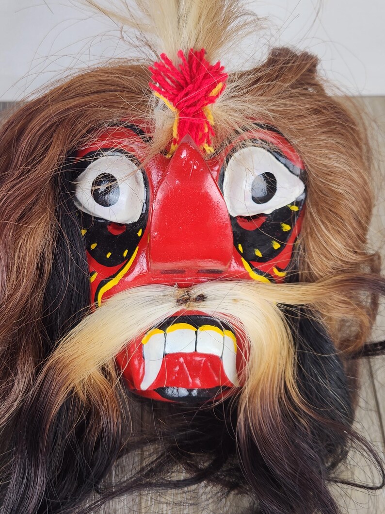 Pu&ograve; includere: Una maschera vibrante e artigianale con un'espressione feroce. La maschera presenta un volto rosso con occhi bianchi, lunghi baffi bianchi e marroni e una bocca rossa con denti bianchi. Capelli castani e neri circondano il volto, con un ciuffo rosso e giallo in cima.