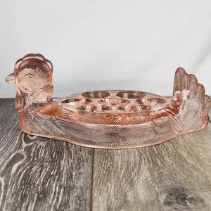 May include: A pink glass hen-shaped egg holder. The hen has a detailed head and tail feathers, with a series of circular indentations for holding eggs. The glass has a translucent quality, and the design is etched with feather details.