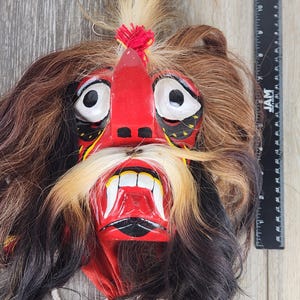 Pu&ograve; includere: Una maschera artigianale con un'espressione feroce. La maschera presenta un volto rosso con occhi bianchi, un naso prominente e una bocca ampia che rivela denti bianchi. &Egrave; adornata da capelli castani, neri e biondi e da un nappa rossa. La maschera &egrave; probabilmente utilizzata per scopi culturali o teatrali.
